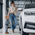 Mujer en el concesionario de autos con su bebé parado cerca de un auto nuevo blanco que financiará con un préstamo para auto de Alianza.