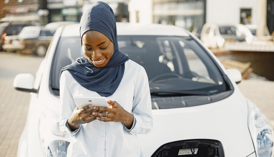 Woman-in-beautiful-blue-head-scarf-is-refinancing-a-car-loan-while-leaning-against-her-car-while-it-is-charging.